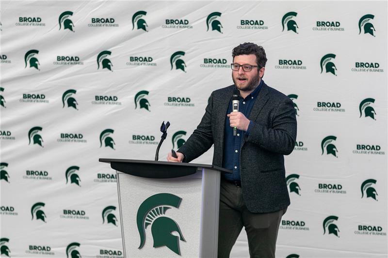 Speaker at a podium with Michigan State University and Broad College of Business backdrop, holding a microphone during a presentation.