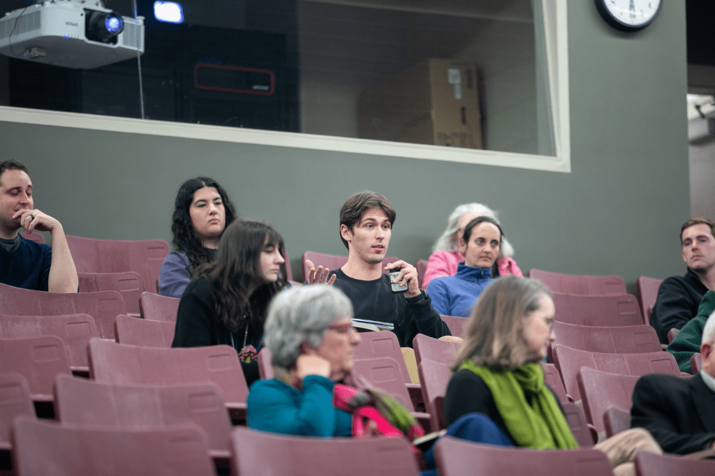 Student in a lecture hall speaks into a microphone during a Q&A session as peers and faculty look on.