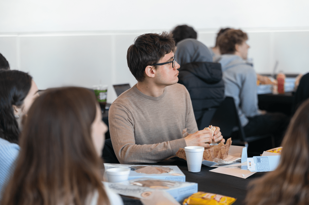 Students seated at round tables eat lunch and listen attentively during a campus event.