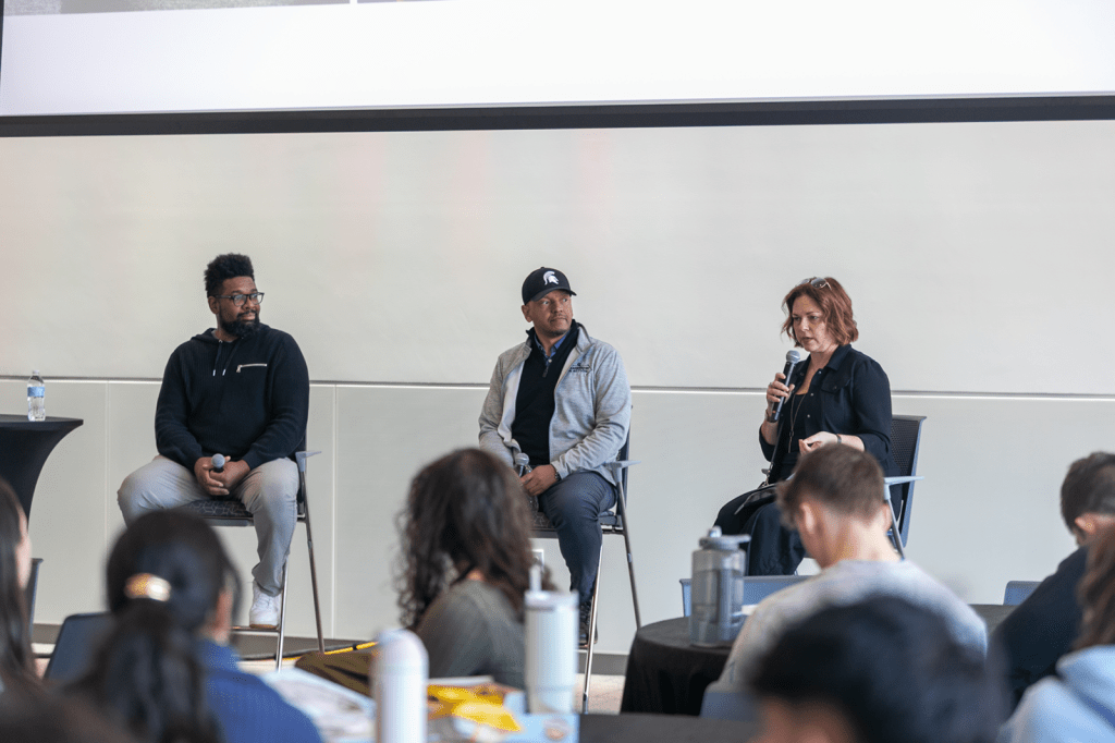 Panel discussion with three speakers seated on stage, one speaking into a microphone while addressing an audience.