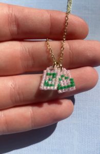 Close-up of a hand holding a delicate gold chain necklace with two small beaded charms, each featuring a green “S” on a light pink background