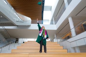 A graduate in a green cap and gown stands on a wide staircase inside a modern building, tossing her cap into the air with one arm raised.