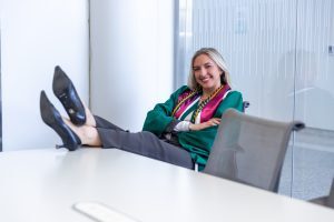 A graduate in a green gown and honor cords leans back in a chair with her feet up on a desk, smiling confidently in a bright, modern interior space.