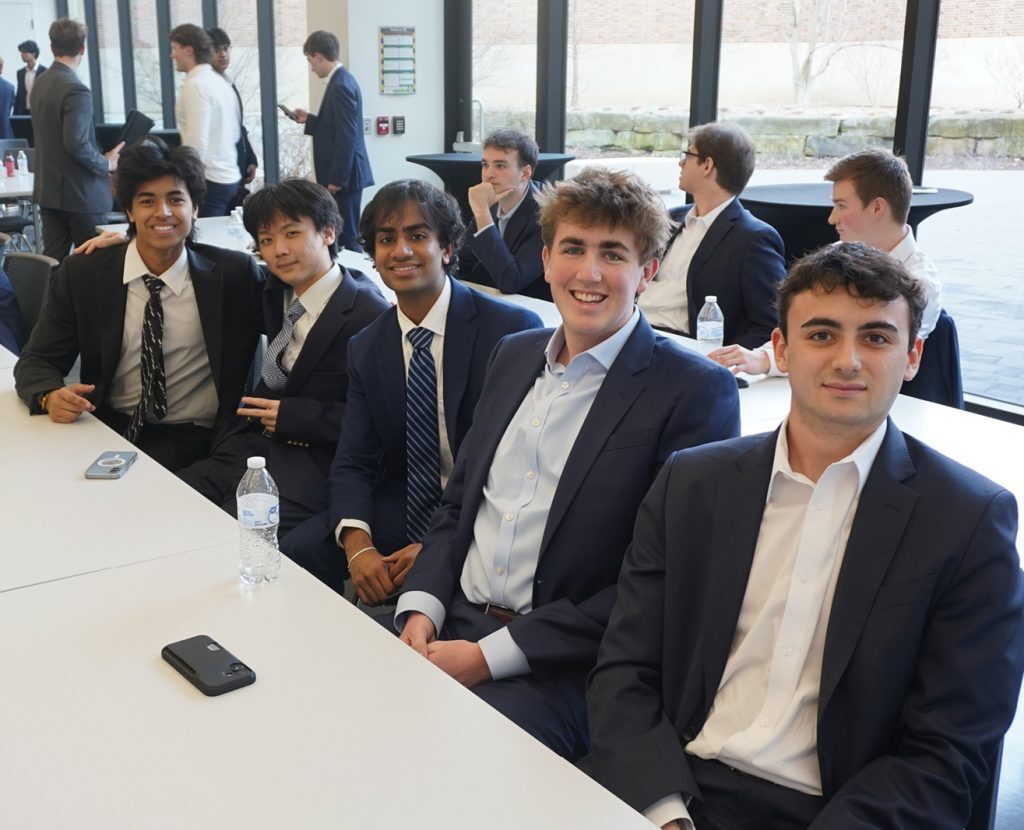 Group of business students in professional attire sit together at a table, smiling at the camera during a networking or competition event.