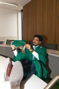 Seated student in a graduation gown hold a tassle on a graduation cap 