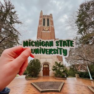 Hand holding a “Michigan State University” sticker in front of Beaumont Tower on campus, with cloudy skies and trees surrounding the landmark.