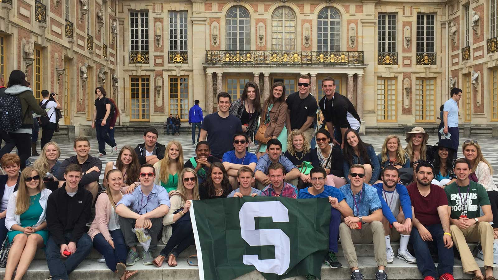 MSU students studying abroad gather for a group photo outside on a set of steps. holding a Michigan State flag.