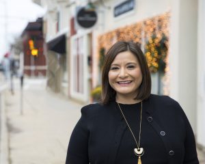 Milinda Ysasi in a black shirt and long silver necklace stands on a beautiful city street and smiles at the camera