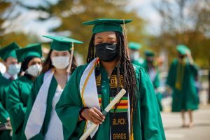 Students at outdoor commencement ceremony, wearing caps, gowns, masks.